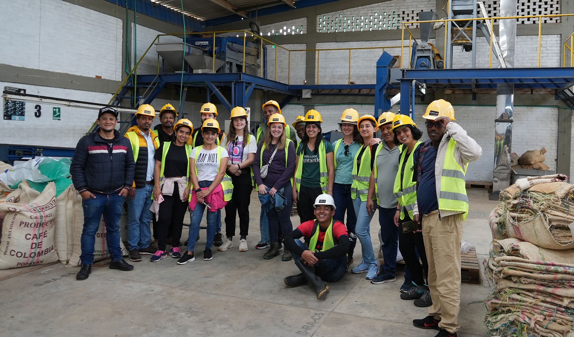 A large group of people wearing yellow hard hats stand together and smile at the camera, whilst stood in a large warehouse with coffee bags either side